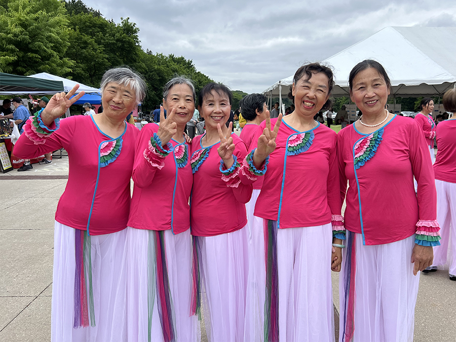 Five smiling Asian America women wear bright pink tops and long pale pink skirts. There are holding up their hands in a peace sign