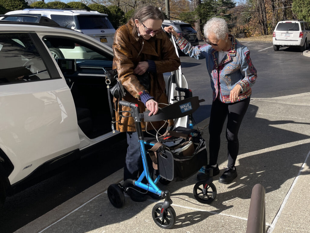 woman helping second woman out of a car and to her walker