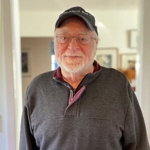 Man with trimmed beard and eyeglasses, baseball hat, faces camera