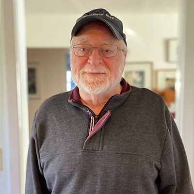 Man with trimmed beard and eyeglasses, baseball hat, faces camera