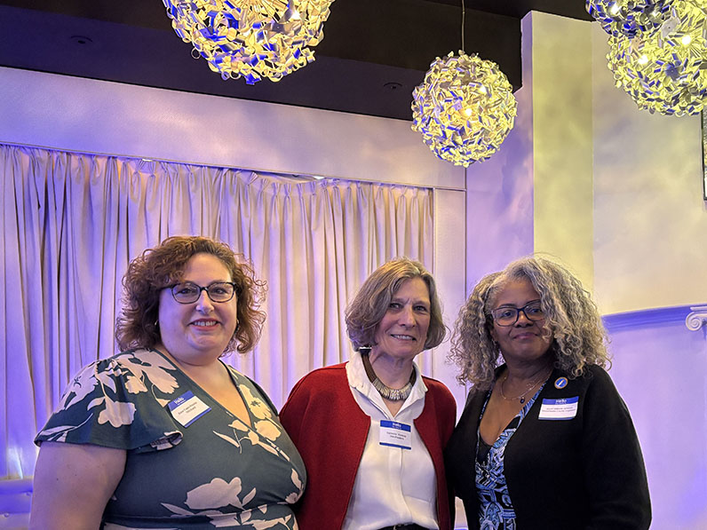 Three standing women stand in front of curtain and below light fixtures