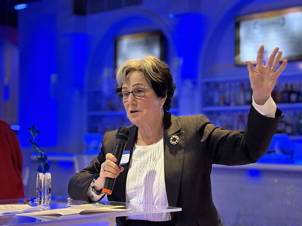 Woman with short dark hair with grey streak raises her left arm as she speaks at podium