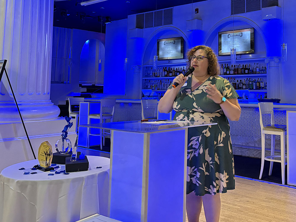 Woman in dark green dress with floral print addresses people from the podium